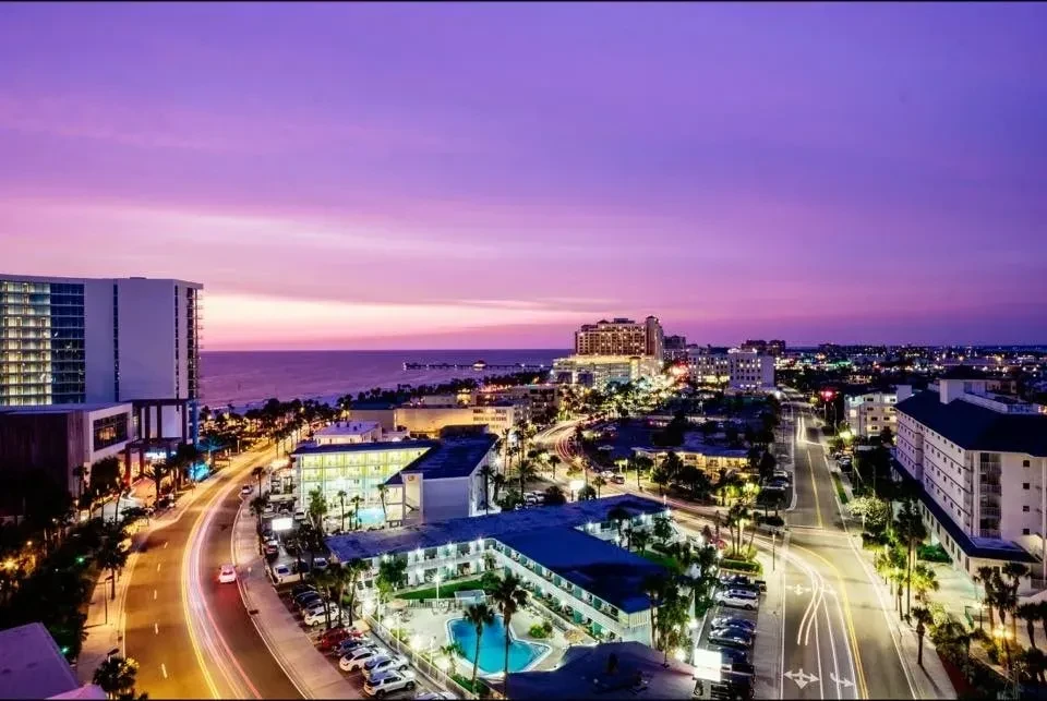 Long-exposure aerial of vibrant coastal city at dusk with light streaks.