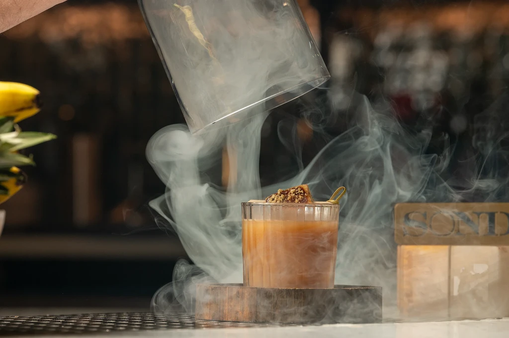 Cocktail with nut brittle garnish, smoke billowing from lifted cloche.