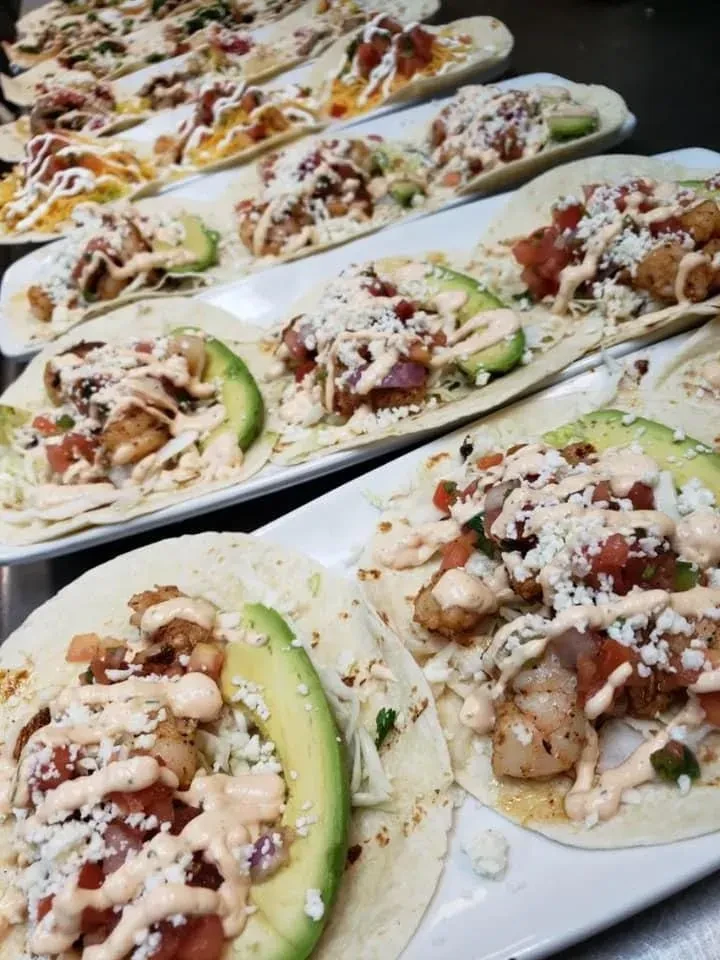 Multiple soft tacos with avocado, cheese, and sauce on white plates.