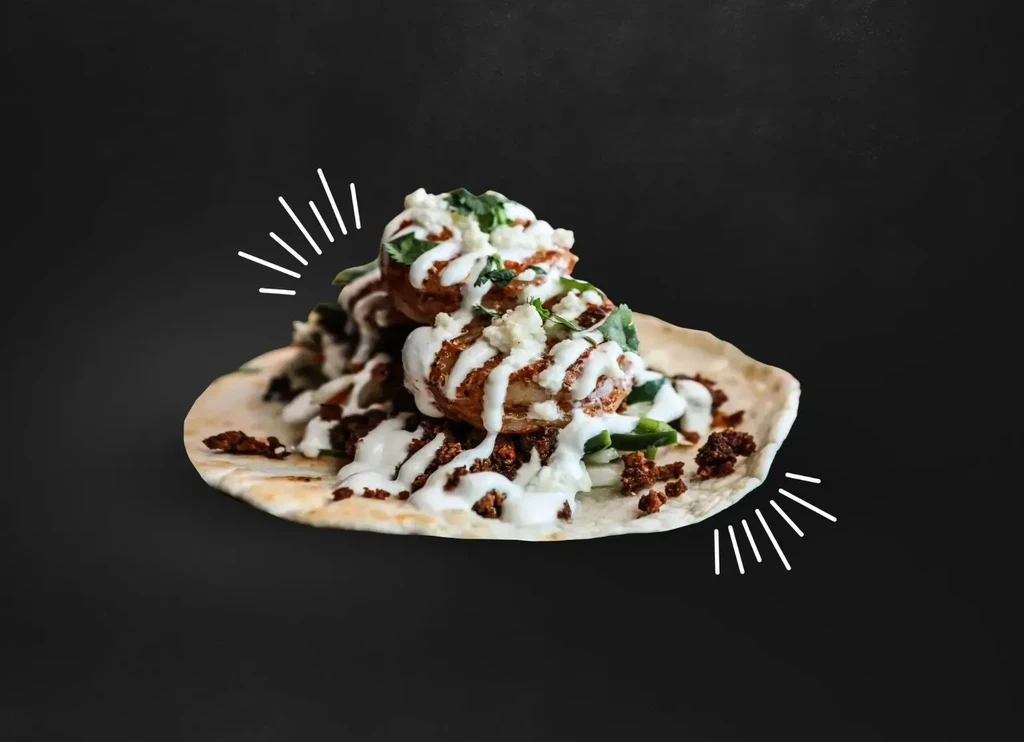 Close-up of a taco with fried protein, white sauce, ground meat, vegetables, cilantro.
