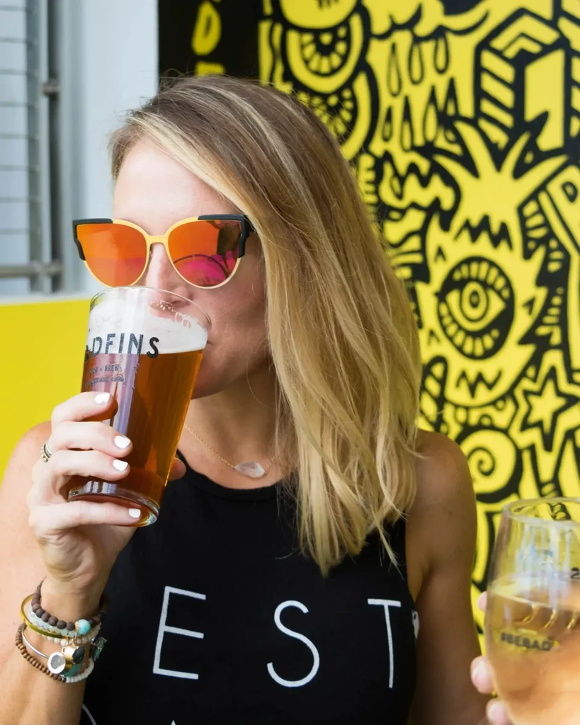 Woman in sunglasses sips beer; vibrant graffiti background.