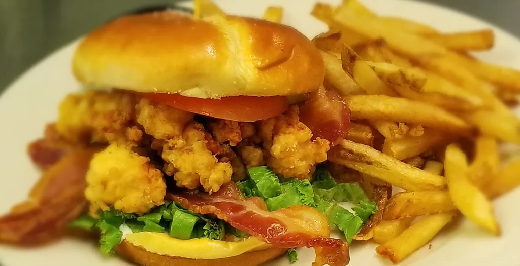 Stacked fried shrimp/chicken sandwich with bacon, lettuce, tomato, and fries.