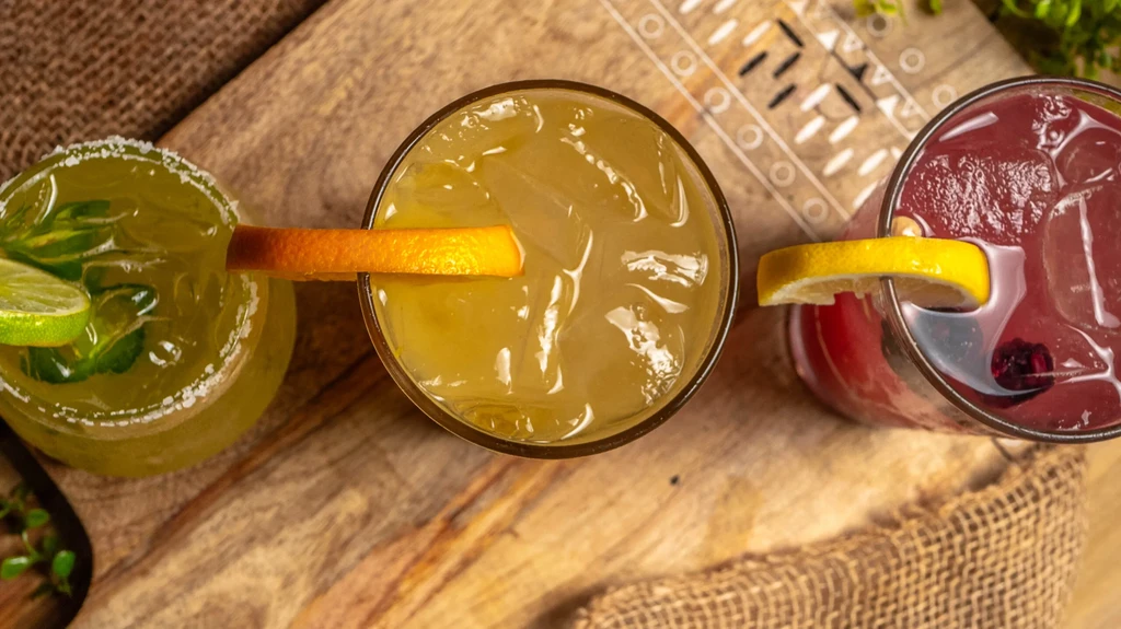Overhead: Three colorful cocktails with garnishes on a rustic board.