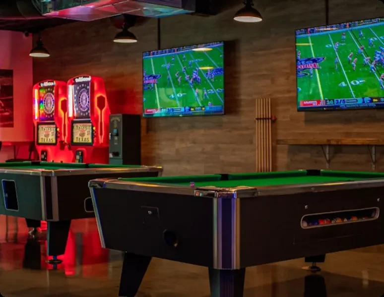 Dimly lit bar with pool tables, red dartboards, and football on TVs.