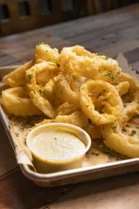 Golden-brown fried rings with herbs and dipping sauce in a tray.