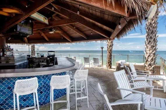 Empty tropical beachfront bar, blue tiled, thatched roof, ocean view.