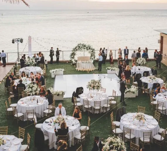 Elegant outdoor wedding reception by water, with guests, tables, floral arch.