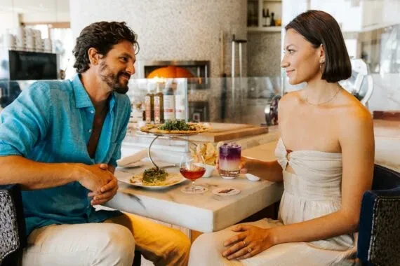 Couple smiling, enjoying food and cocktails at modern restaurant with pizza oven.