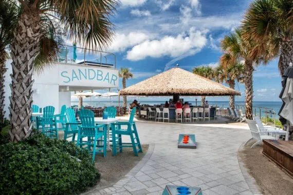 SANDBAR outdoor beach bar, thatched roof, seating, and ocean backdrop.