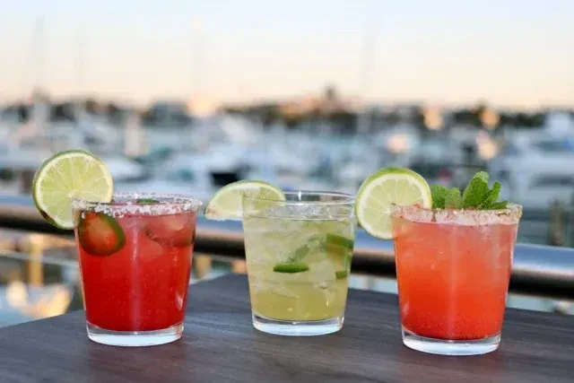Three colorful garnished drinks in clear glasses, blurred marina background.