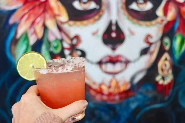 Hand holds pink cocktail with lime, blurred sugar skull mural background.