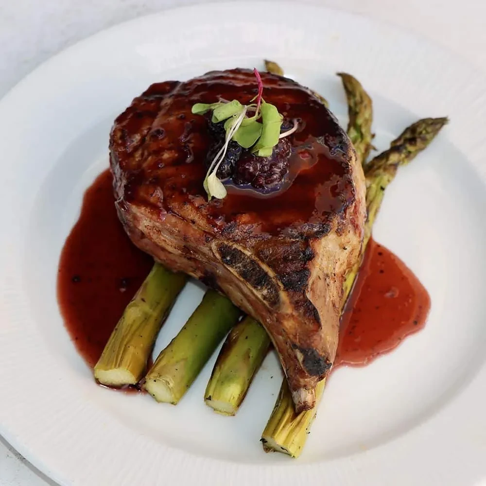 Plated bone-in chop with dark red sauce, asparagus, microgreens, and berry.