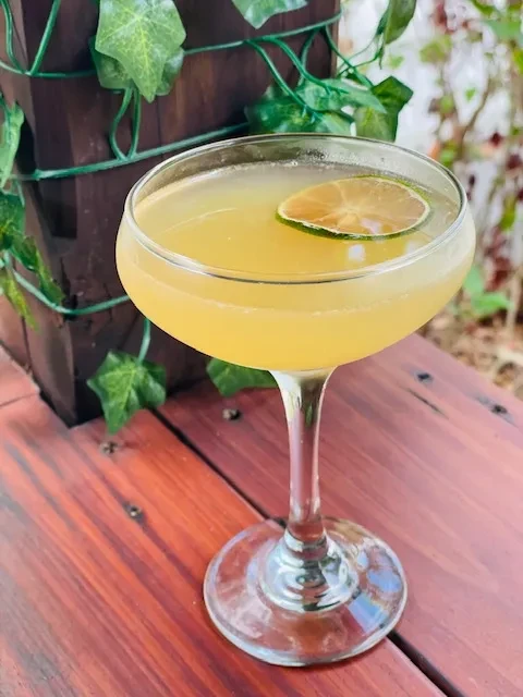 Amber cocktail with lime slice in coupe glass on wooden table.