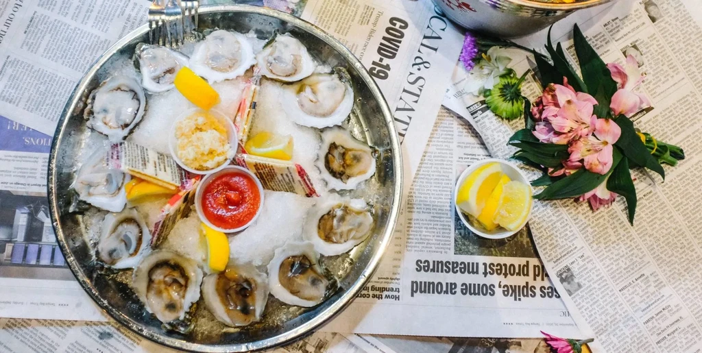 Raw oysters on ice, with lemon, sauces, and flowers on newspaper.