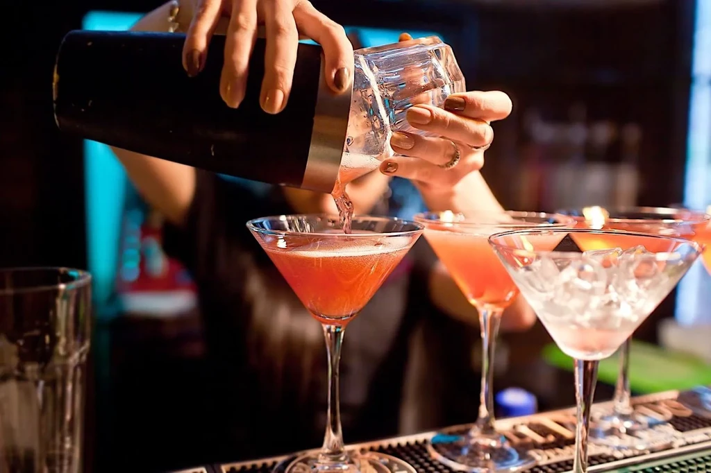 Hands pour a pink cocktail from a shaker into a martini glass.