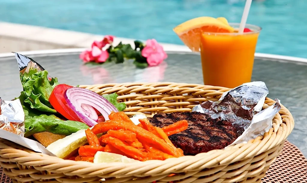 Wicker basket holds a grilled burger, fries, salad, and drink poolside.