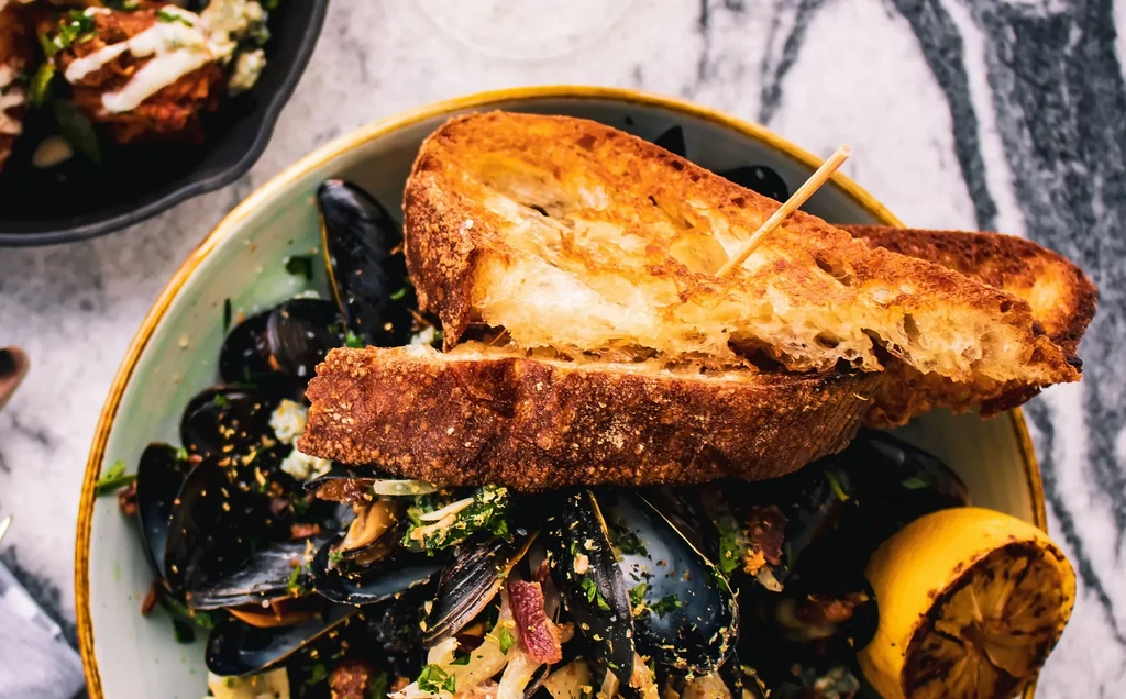 Mussels with grilled bread, herbs, and charred lemon in a bowl.