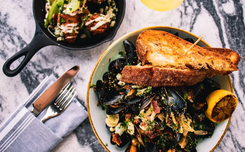 Overhead: mussels with toast, crispy chicken, and drink on marble table.