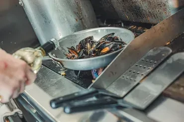 Professional kitchen: Hand pours wine into pan of cooking mussels.