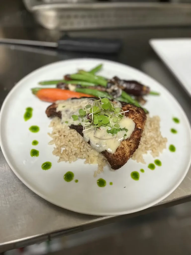 Plated golden-brown cutlet, rice, creamy sauce, vegetables, and microgreens.