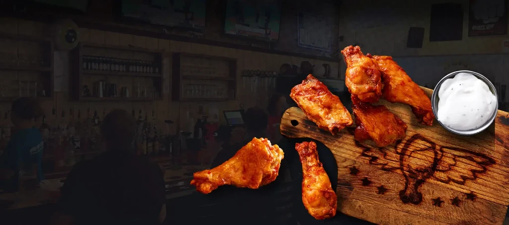 Cooked chicken wings and drumsticks on a board, white sauce, bar background.