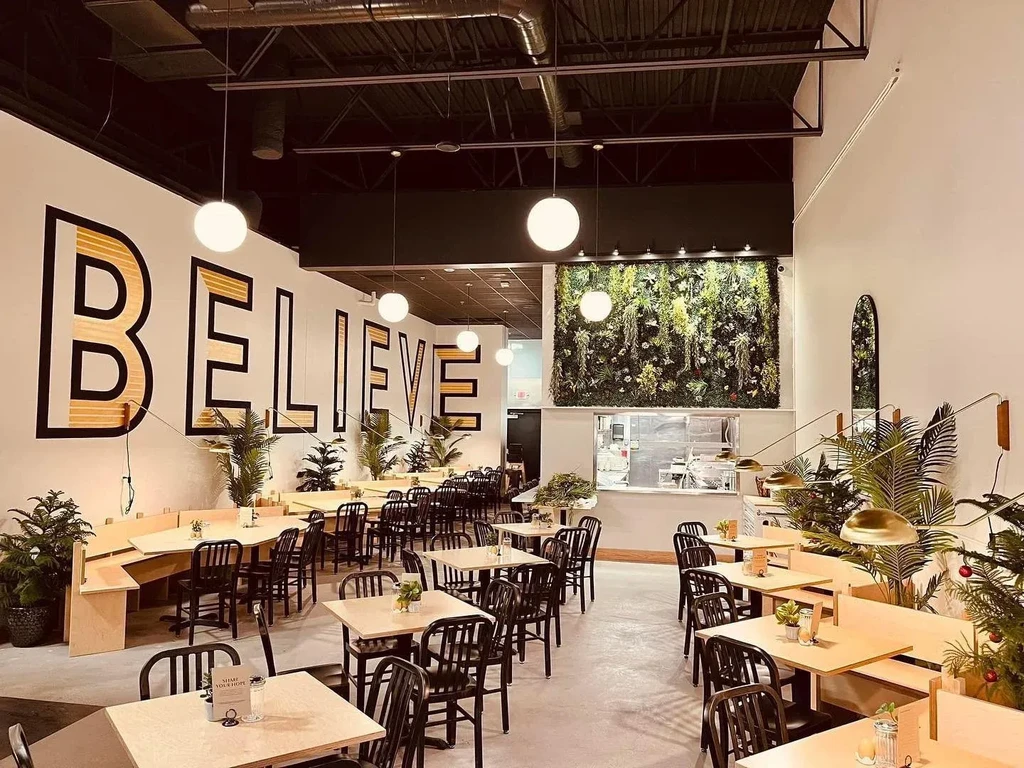 Modern restaurant with "BELIEVE" mural, wood tables, and living wall.