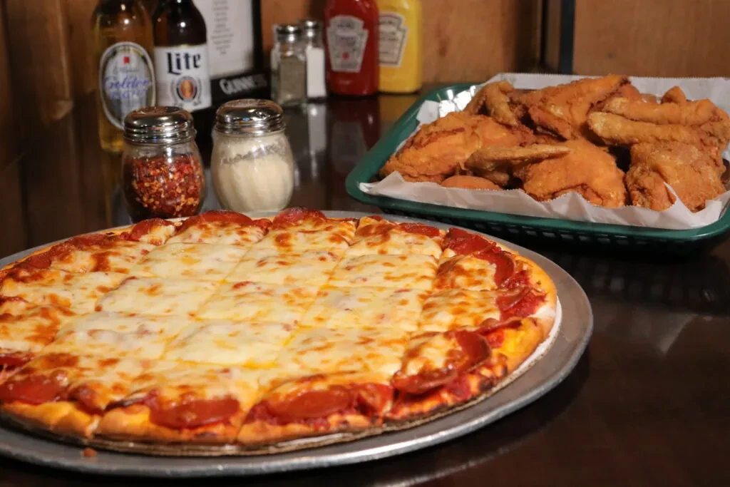 Large pepperoni pizza, fried chicken, condiments, and beer on table.