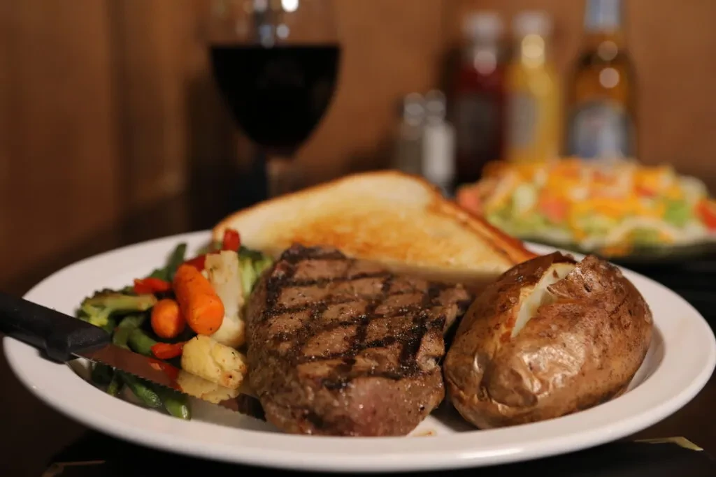 Close-up of grilled steak, baked potato, vegetables, and bread on a plate.