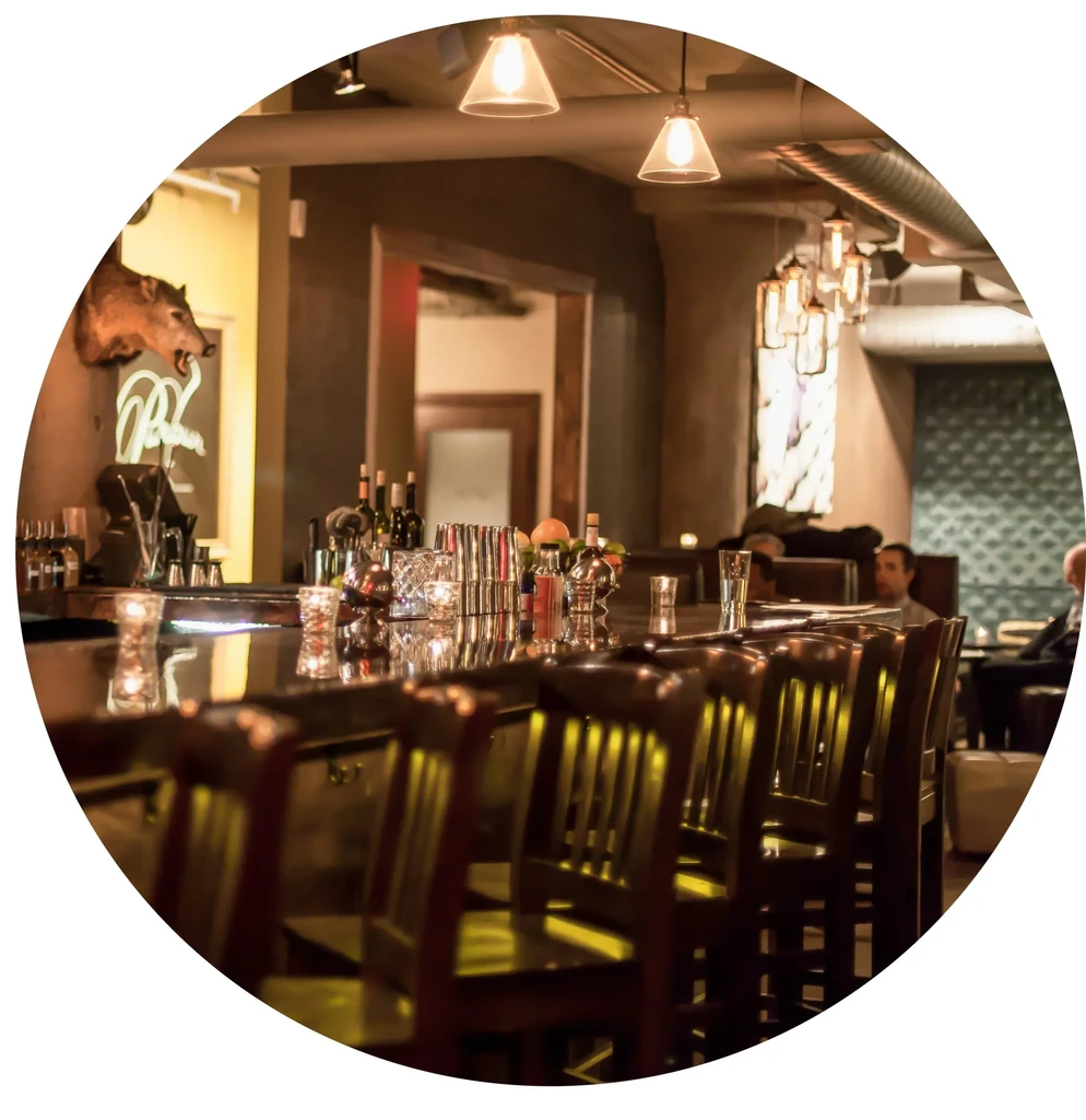 Dimly lit bar interior, reflective counter, stools, bottles, and pendant lights.