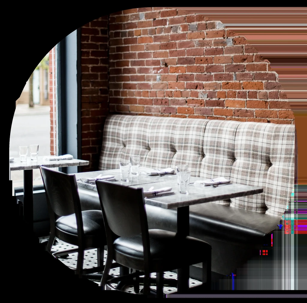 Empty, well-lit restaurant dining area with tables, plaid banquette, brick wall.