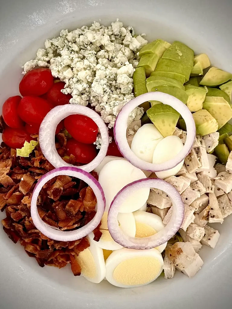 Deconstructed salad in white bowl: chicken, egg, avocado, bacon, cheese, tomatoes.