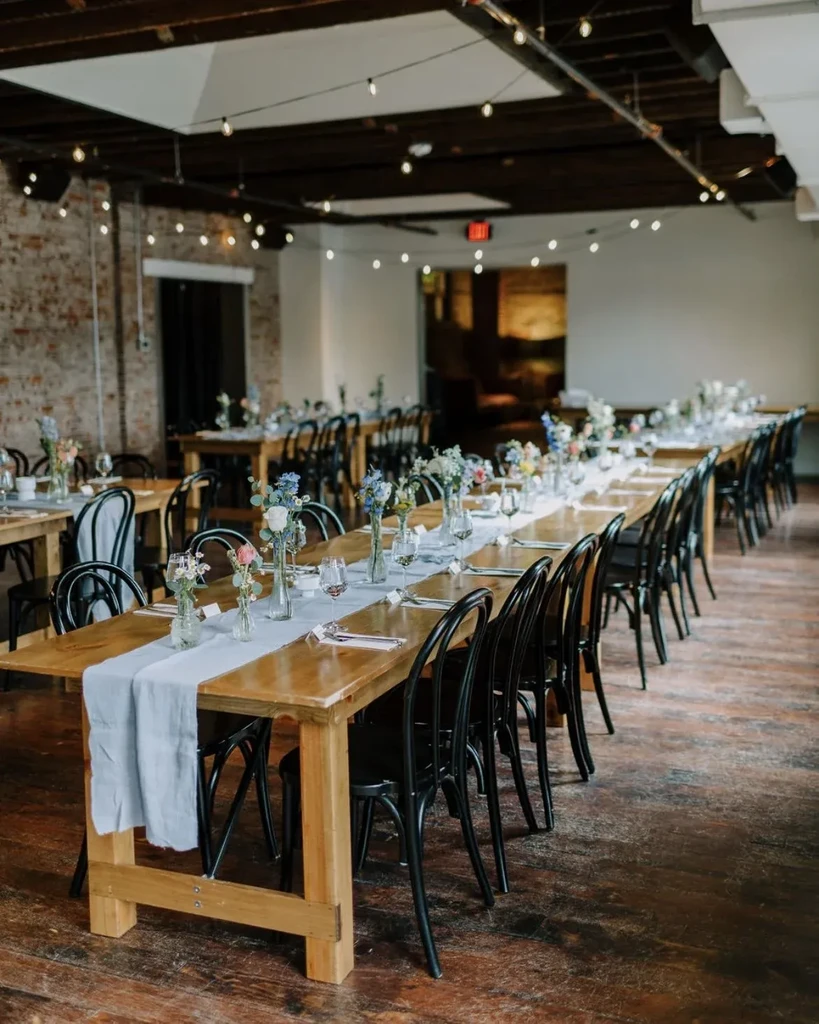 Empty rustic event room with long tables, floral centerpieces, and string lights.