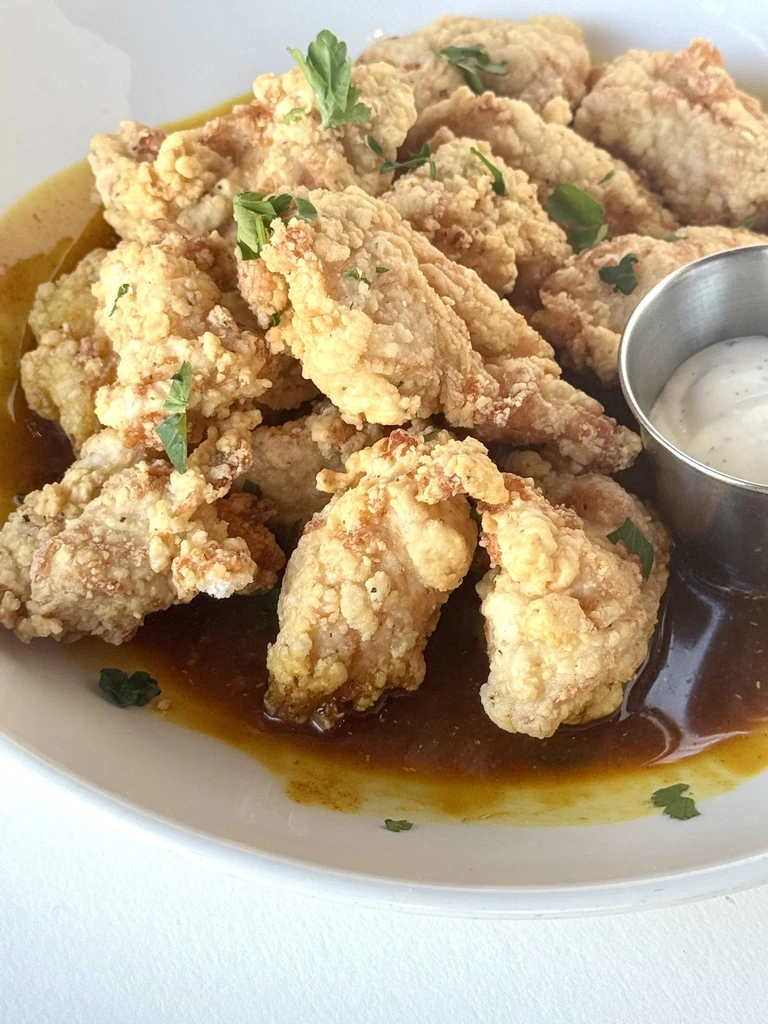 Plate of golden fried chicken tenders in dark sauce, with herbs and white dip.