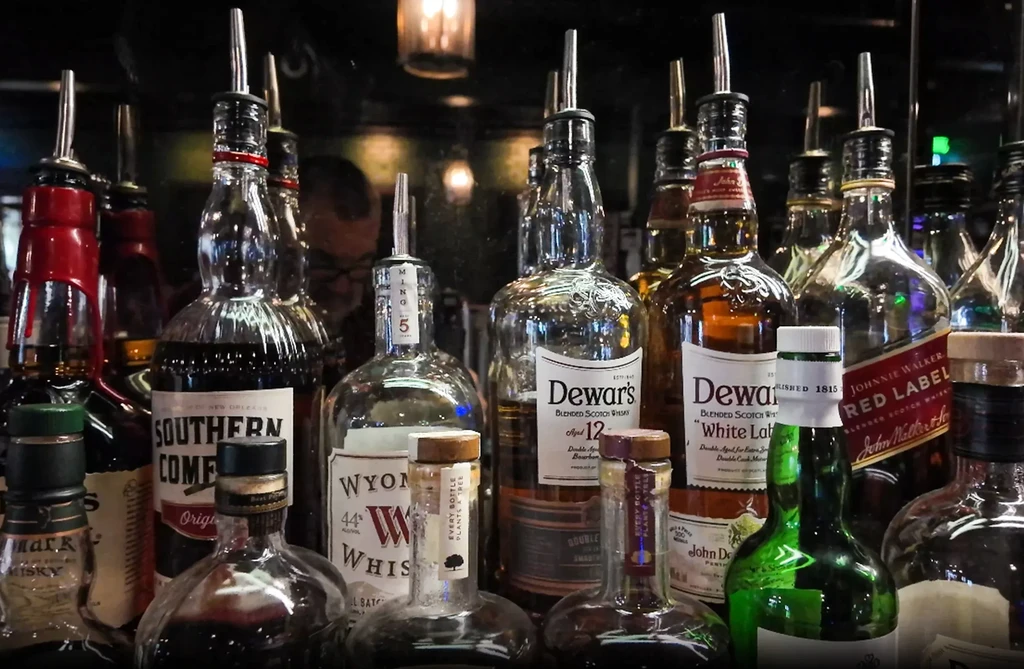 Labeled whiskey bottles with pour spouts on a dimly lit bar shelf.