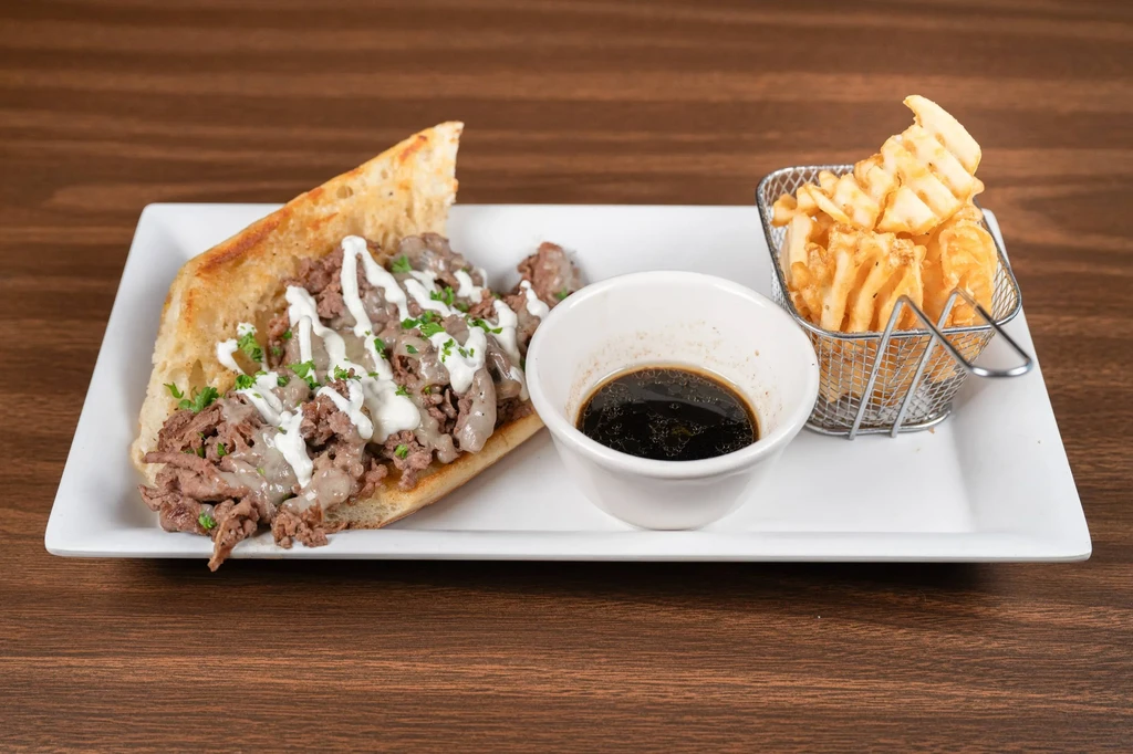 Open-faced cheesesteak, waffle fries, and au jus on a wooden table.