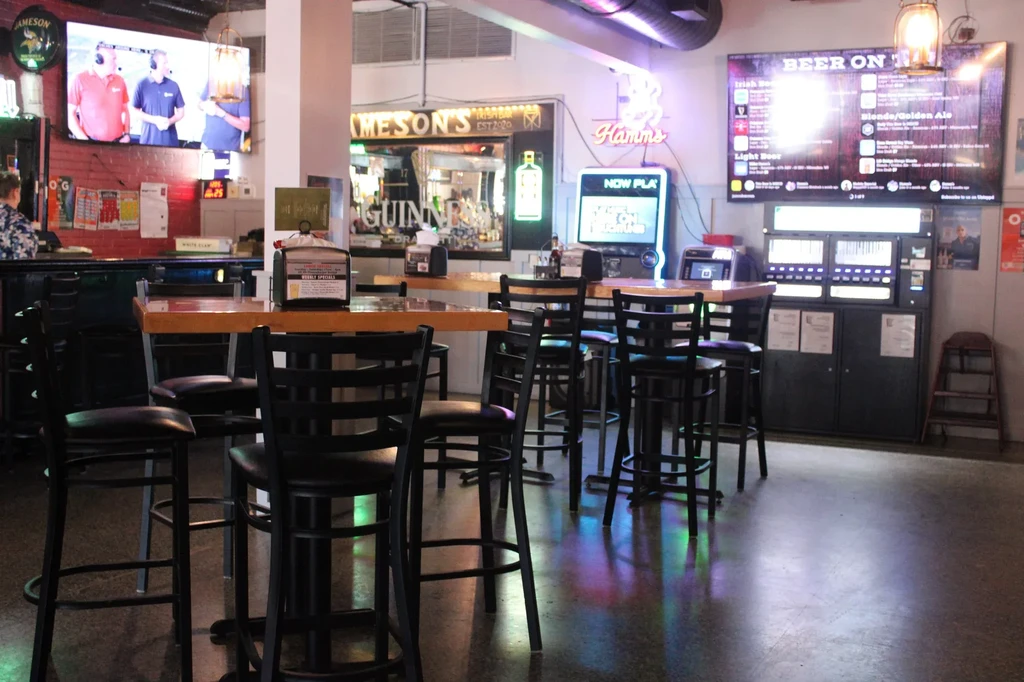 Dimly lit bar with high-top seating, multiple screens, and neon signs.