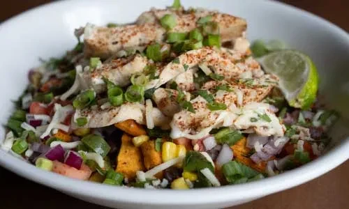 A colorful chicken and vegetable bowl with corn, cheese, and lime.