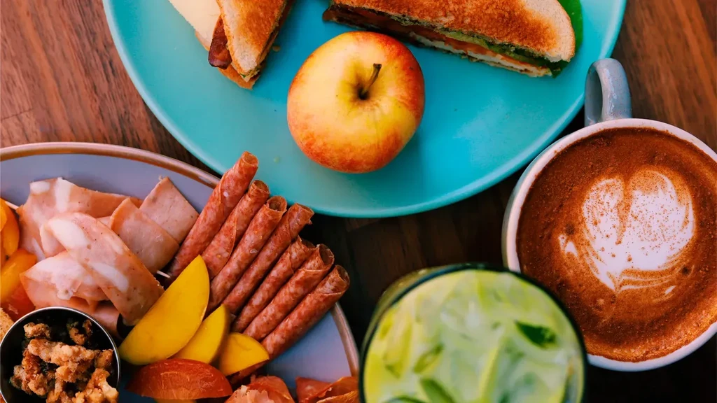 Overhead: sandwiches, charcuterie, coffee, and icy green drink on wooden table.