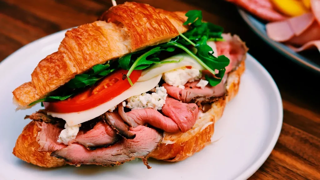 Sliced croissant sandwich with roast beef, cheeses, tomato, and arugula.