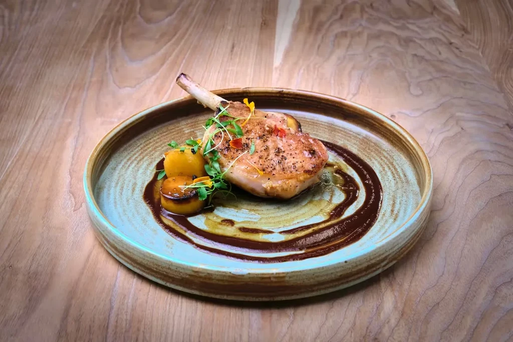 Plated bone-in pork, roasted peaches, microgreens, and flowers with sauce.