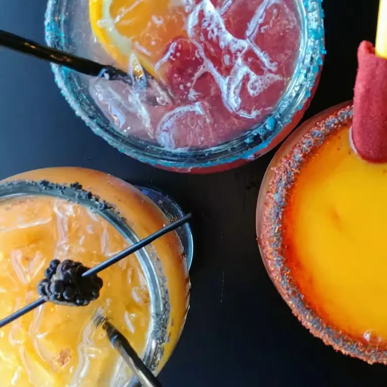 Three colorful drinks with unique rims and garnishes on a dark surface.