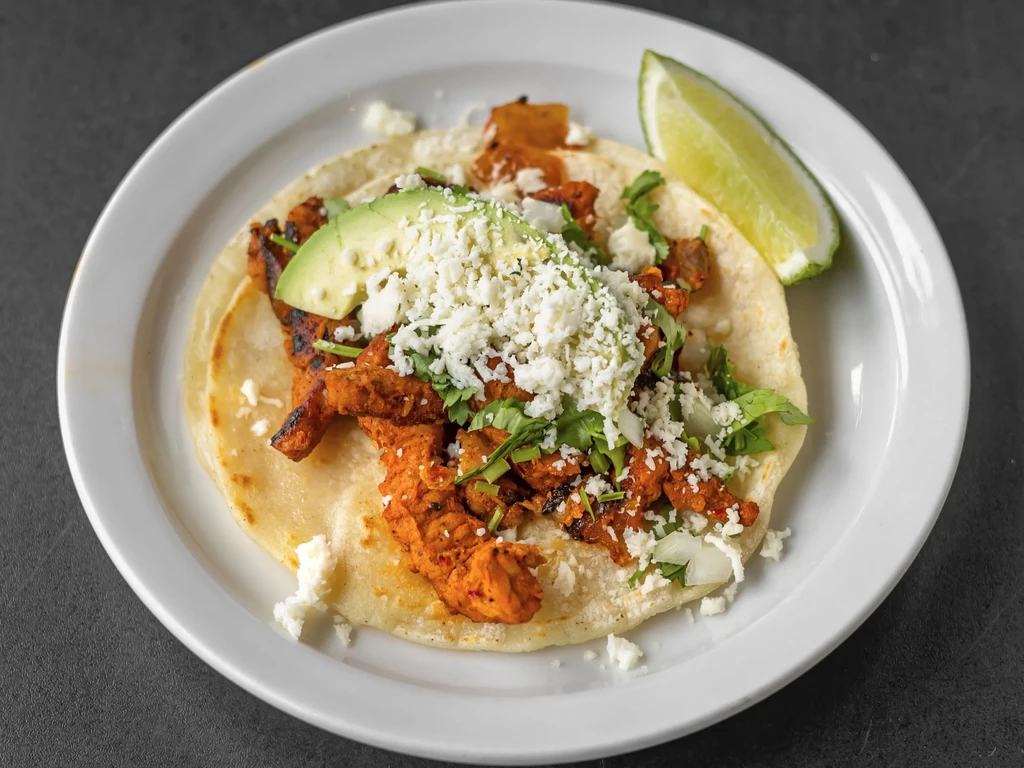 Single taco with meat, cheese, cilantro, avocado, and a lime wedge.