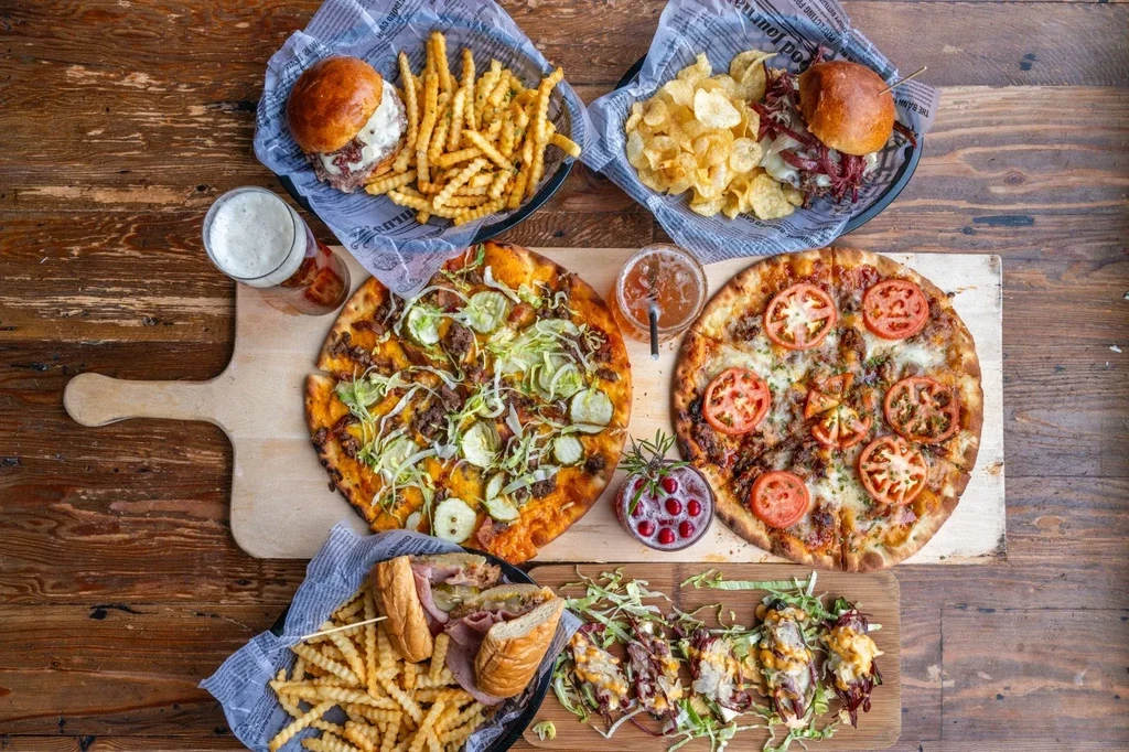 Flat-lay of pizzas, burgers, sub, appetizers, and drinks on a wooden table.