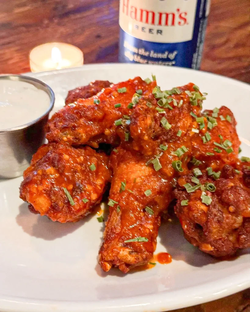 Plate of reddish-orange chicken wings with chives, dip, and beer.