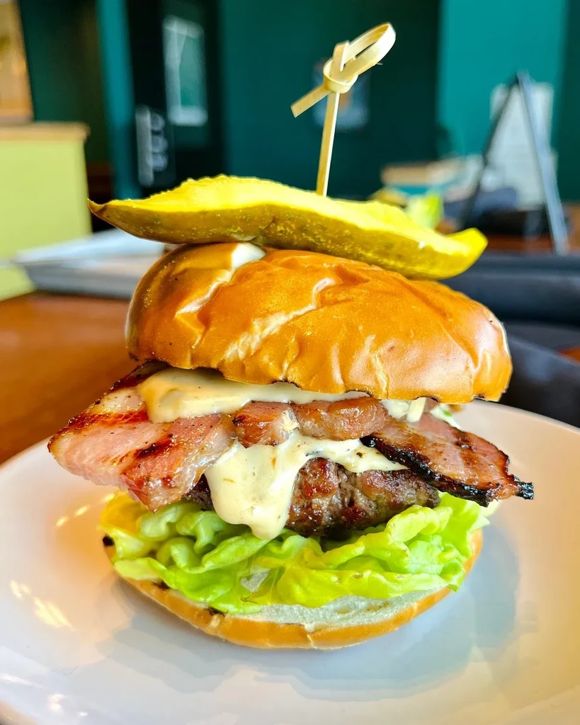 Gourmet burger with thick beef patty, bacon, pickle, and white sauce.