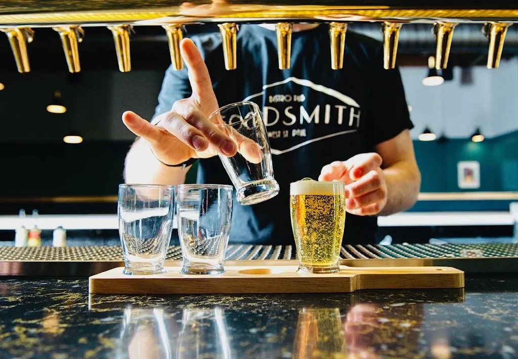 Server arranges beer flight: one golden beer, two empty glasses.