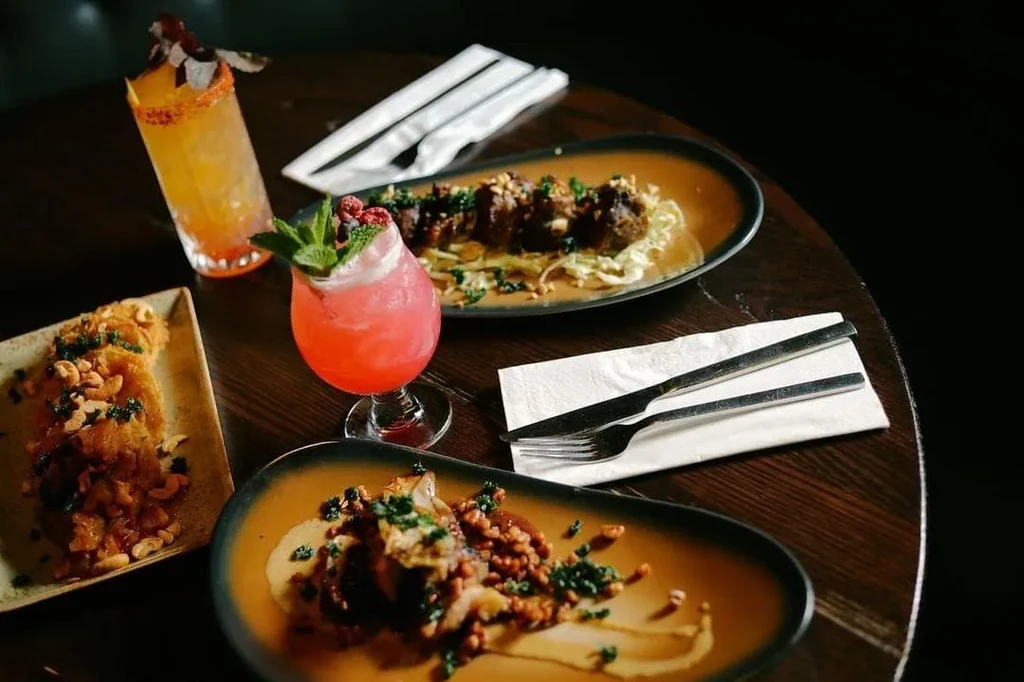 A spread of diverse gourmet dishes and two colorful cocktails on a dark restaurant table.