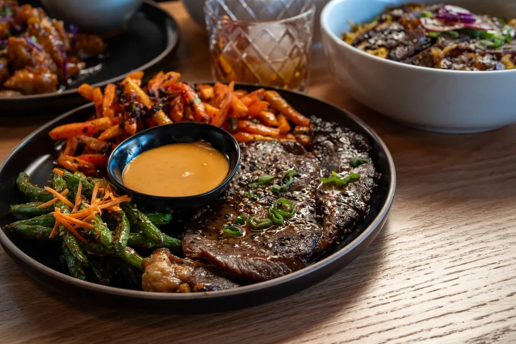 Dark plate with glazed steak, orange fries, green beans, and sauce.