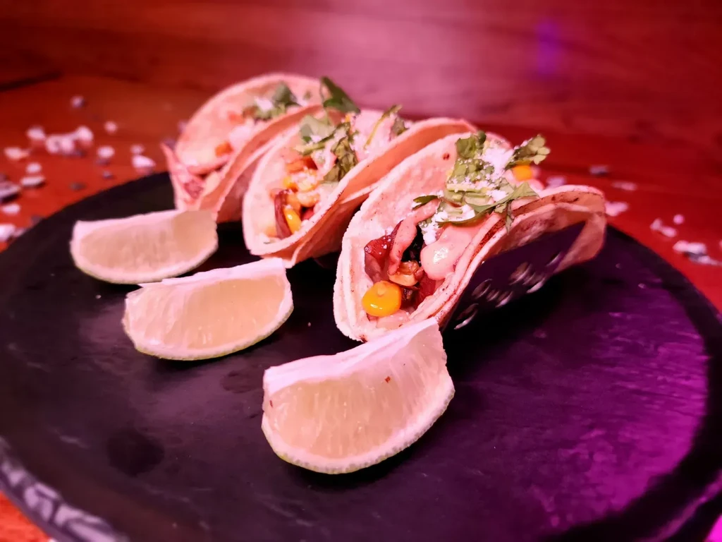 Four garnished tacos and lime wedges on a dark plate.