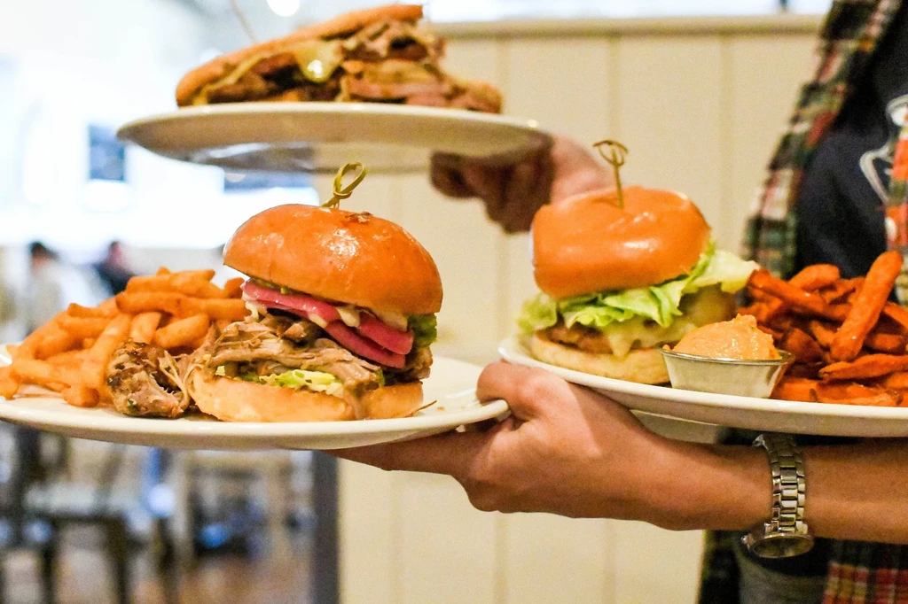 Hands serving multiple plates of sandwiches, French and sweet potato fries.